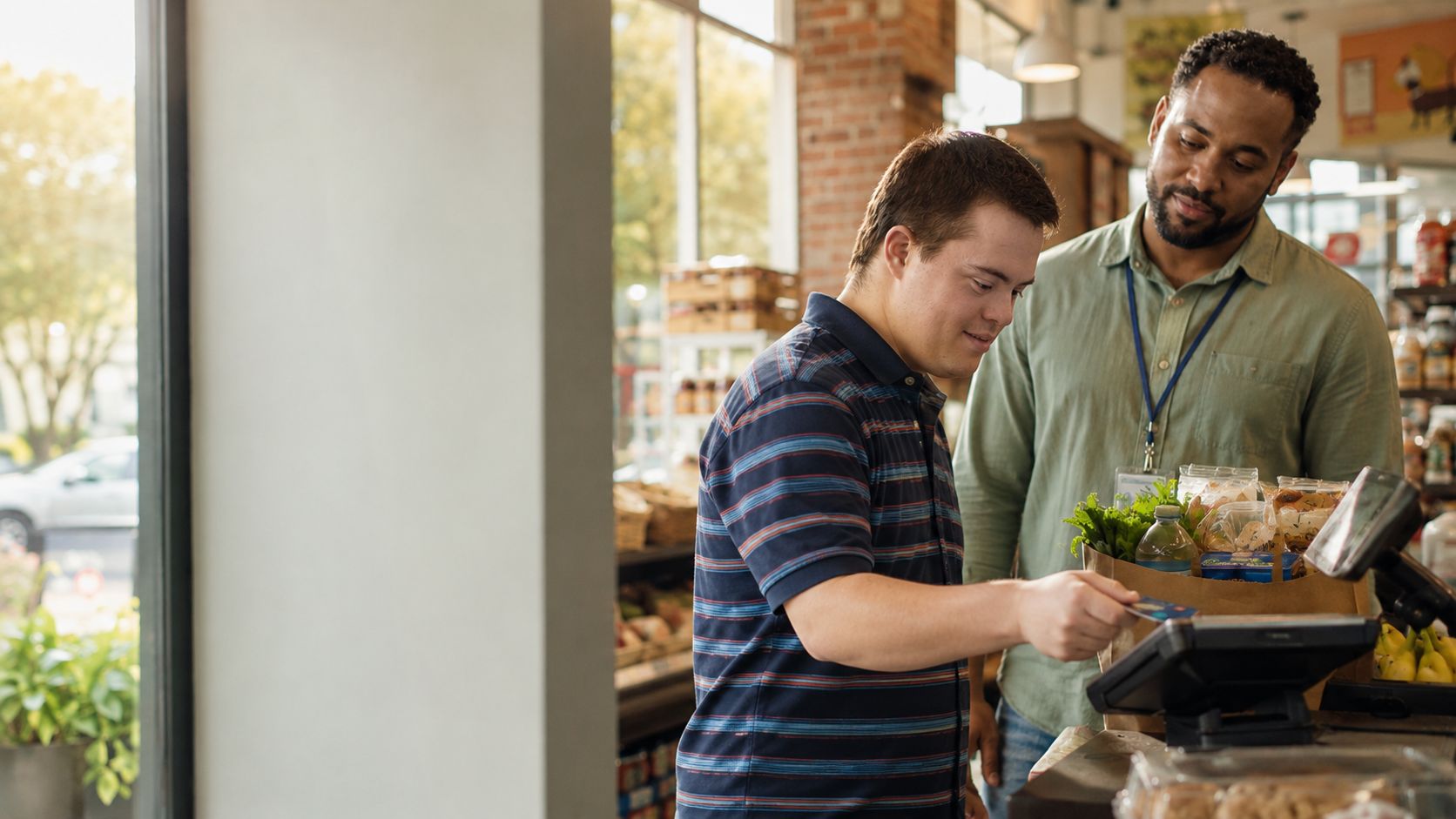 A young man paying for his own groceries at a store counter while a Birch support worker stands alongside him.
