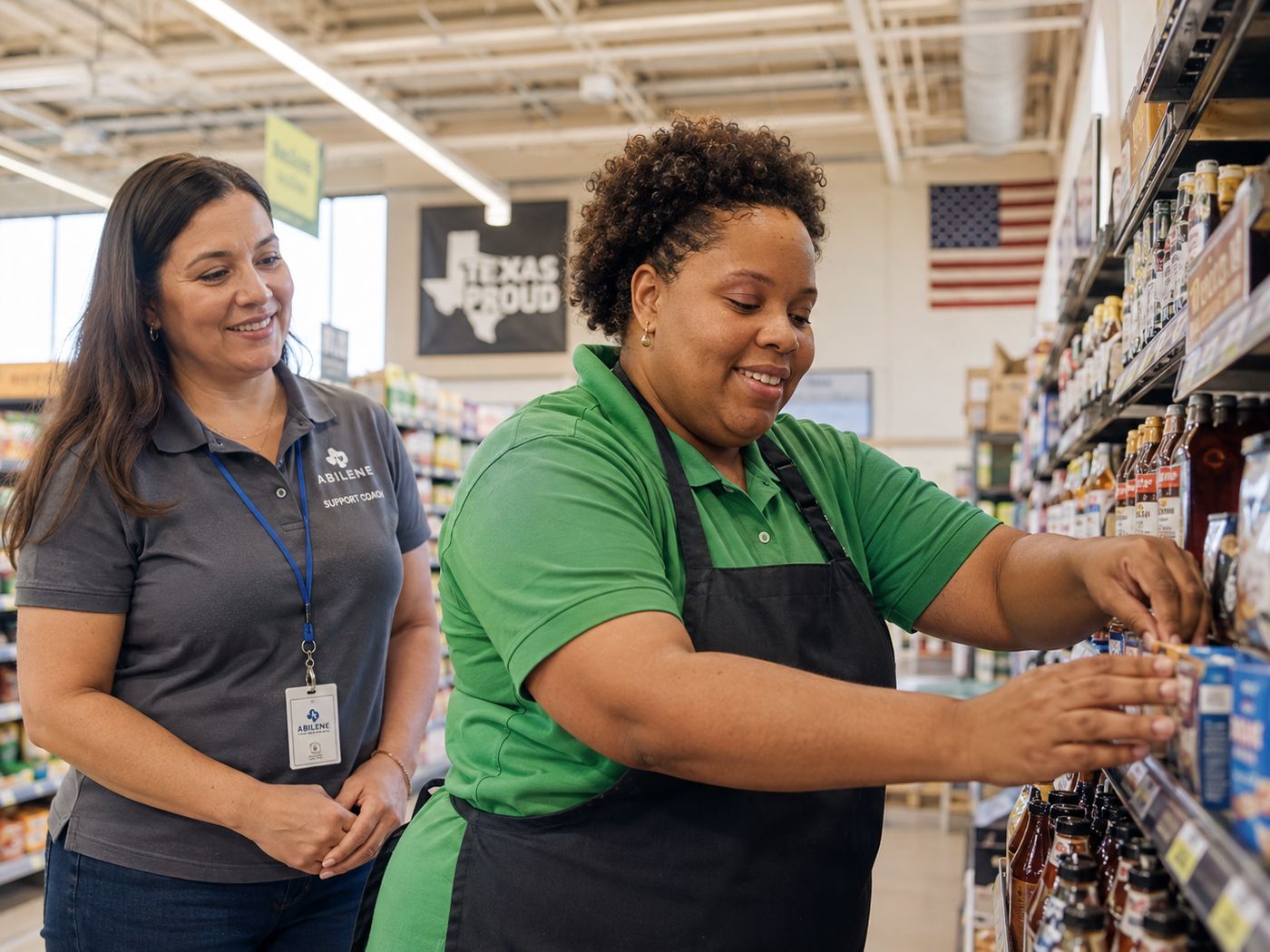 A woman in a supermarket uniform facing up shelves while her support coach stands nearby.